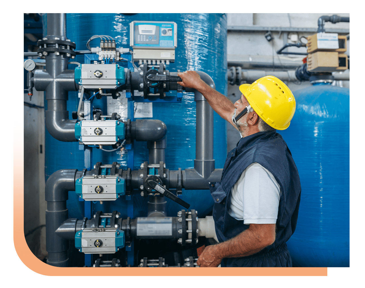 Maintenance worker in a yellow hard hat adjusts a valve on a dense system of blue pipes and equipment in an industrial setting.