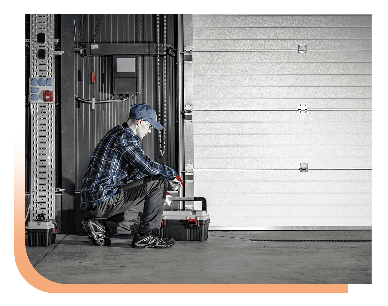 Male technician kneels by a tool box and repairs a large white roll-up garage door in an industrial setting, wearing a blue cap and plaid shirt with gloves