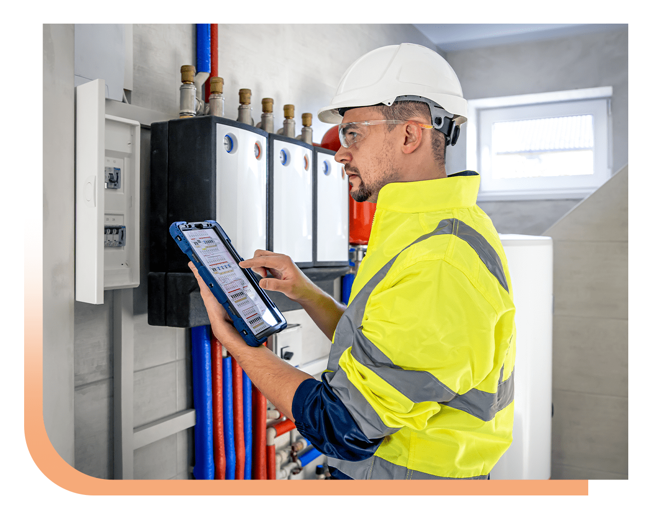 Technician in a yellow safety jacket and white hard hat using a tablet next to a control panel with pipes in an industrial room