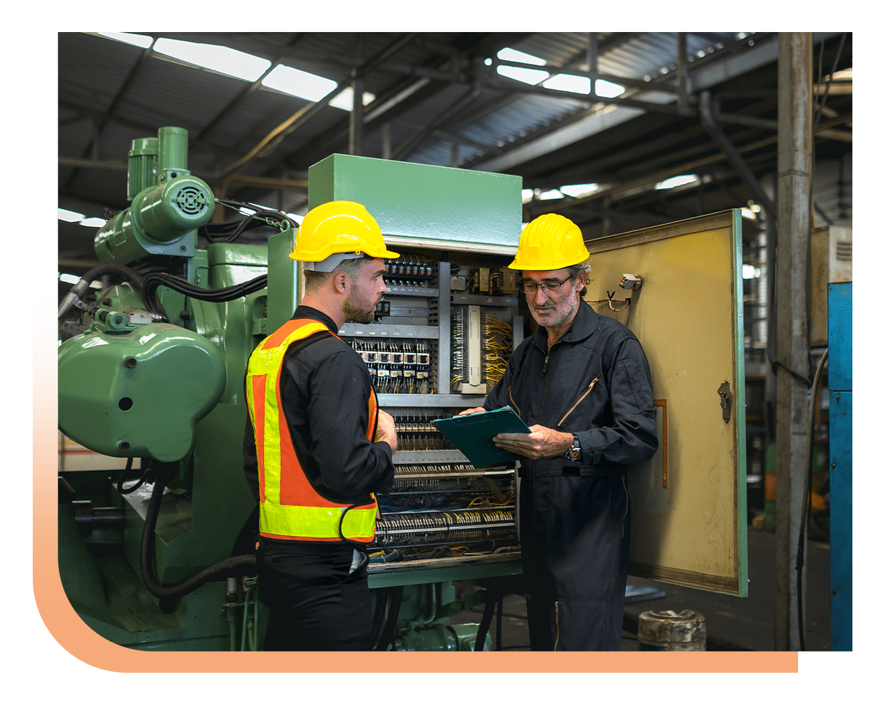 Two factory workers in yellow hard hats inspect an open electrical control panel together, one with a clipboard and safety vest in a large green machine room.