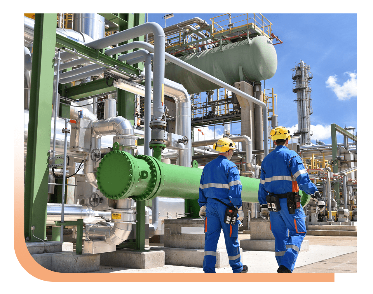 Two workers in blue coveralls and yellow hard hats walk beside a large green industrial pipe network in a plant.