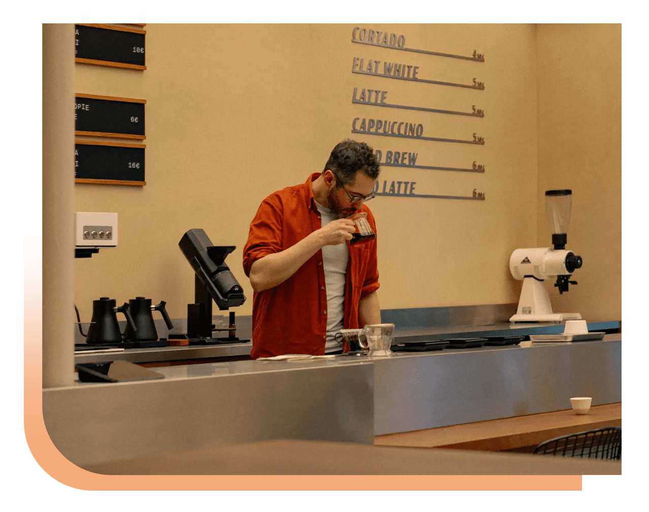 Barista in an orange shirt samples a cup of coffee behind a cafe counter with an espresso machine and grinder, menu boards on the wall behind him